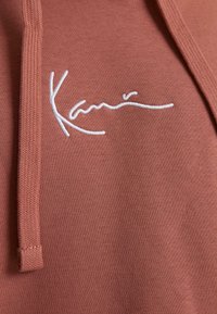 Amberfarvet hættetrøje med en tekstureret overflade, der har hvid broderet branding. Snorejusteringer og ribbede manchetter er synlige.