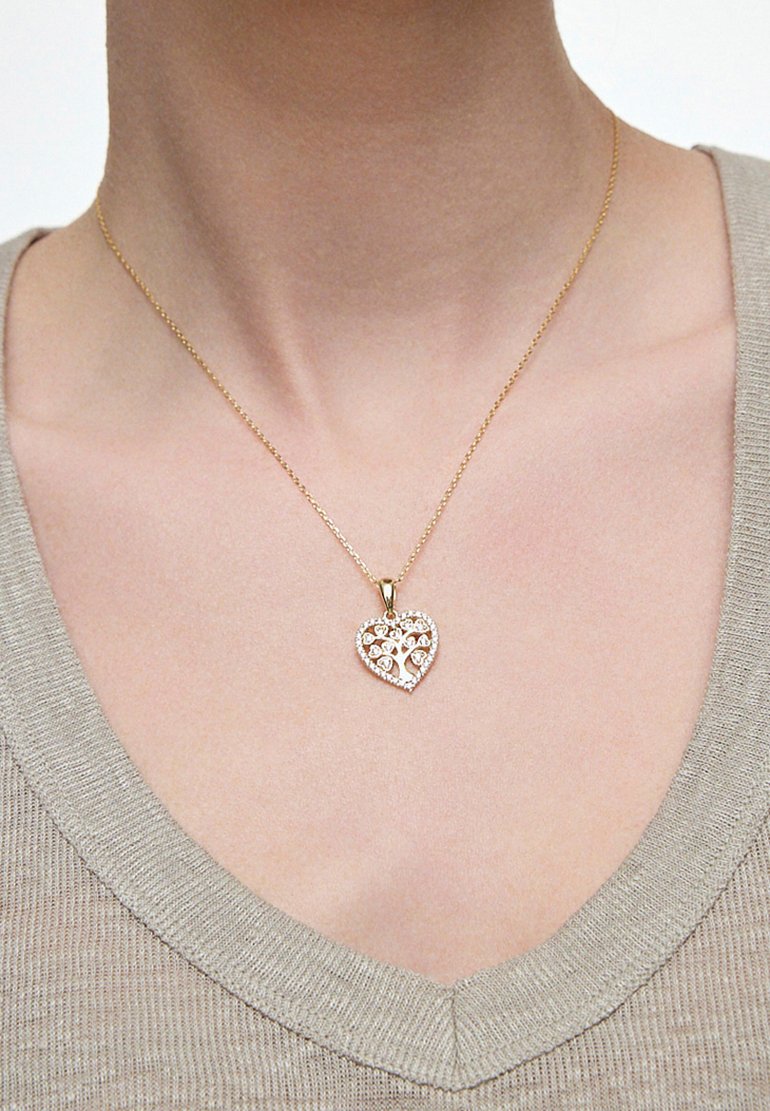 Złoty naszyjnik z sercowym wisiorkiem, ozdobiony przezroczystymi kamieniami szlachetnymi w kształcie drzewa, noszony na jasnoszarym swetrze.