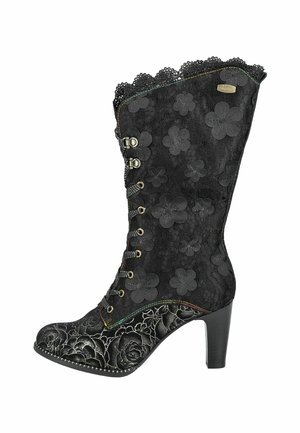 Bottine noire avec un superposé de dentelle florale, bord supérieur festonné et motifs floraux en relief. Dotée d'un talon haut et de détails de couture décoratifs.