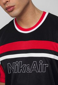Svart tränings T-shirt med röda och vita ränder, med "NikeAir"-logotyp i fet brodyr på framsidan. Rund halsringning.