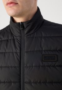 Casaco acolchoado preto com gola alta, apresentando costura horizontal, um fecho éclair preto e um emblema texturizado no peito.