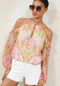 Blusa trasparente a fiori con spalle scoperte, intricate fantasie in rosa e giallo e maniche arricciate. Abbinata a pantaloni corti bianchi.