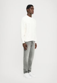 Vit kabelstickad tröja med diamantmönster, i kombination med grå denimjeans och vita sneakers. Modellen står på sidan mot en enkel bakgrund.