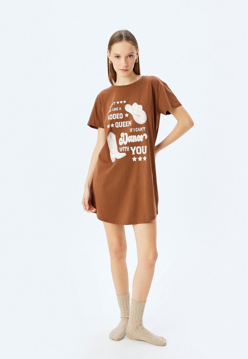 Vestido de camiseta de algodón marrón con estampado gráfico y texto blanco, combinado con botas vaqueras. Mangas cortas y corte holgado. Calcetines de canalé color beige.
