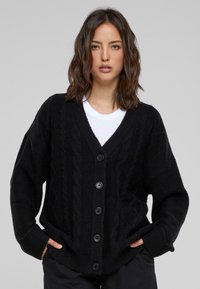 Zwarte gebreide cardigan met een kabelpatroon, oversized pasvorm, V-hals en zes grote knopen. Geribde manchetten en zoom, met een zachte textuur.