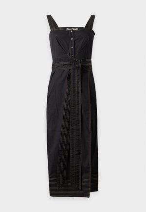 Donna che indossa un abito midi nero senza maniche con corpetto abbottonato, cintura annodata in vita e tacchi neri a punta su sfondo bianco.