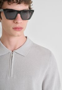 Mlad čovjek nosi crne oversize sunčane naočale i svijetlosivi pleteni pulover sa zip zatvaračem nasuprot jednostavne sivoj pozadini.