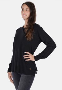 Maglione nero lavorato a maglia con maniche lunghe, scollo arrotondato, orlo a coste e spacchi laterali. Presenta un piccolo logo metallico in basso a sinistra.