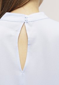 Blusa blu chiaro con tessuto testurizzato, caratterizzata da un'apertura a oblò sul retro e due chiusure a bottone sul collo.