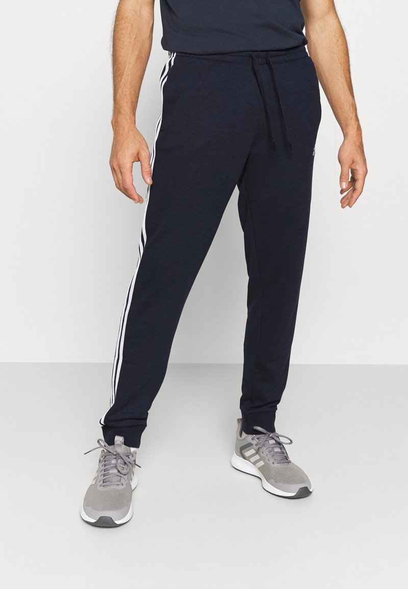 Homme portant un pantalon de jogging bleu marine avec des bandes blanches sur les côtés et des chaussures de sport grises, debout sur un sol gris clair contre un mur blanc.