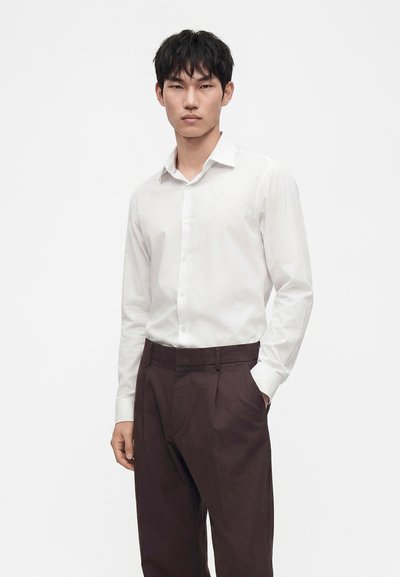 Jeune homme portant une chemise blanche boutonnée et un pantalon marron foncé, debout avec une main dans la poche, sur un fond uni.
