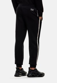 Hombre con pantalones de chándal EA7 negros con franjas laterales blancas y zapatillas negras, de pie contra un fondo claro y liso.