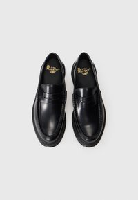Par de mocassins de couro preto brilhante da Dr. Martens com solas grossas, detalhe de costura no topo e palmilhas com logótipo em fundo branco liso.