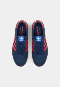 Blå ruskind sneakers med røde striber, med et marineblåt snørebåndsdesign og et blåt logo på tungen. Gummisålen er synlig.