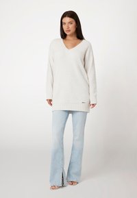 Witte gebreide trui met een V-hals en lange mouwen, met een gestructureerd patroon. Gecombineerd met lichtblauwe flared jeans en open schoenen.