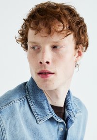 Giacca di jeans azzurro chiaro con colletto, caratterizzata da cuciture visibili e una texture consumata; indossata sopra una camicia marrone, che mette in evidenza dei riccioli di capelli rossi.