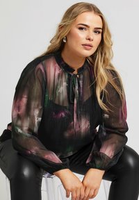 Camicia nera trasparente con motivi floreali in rosa e verde, caratterizzata da un colletto con volant, dettaglio con fiocco e polsini elasticizzati.