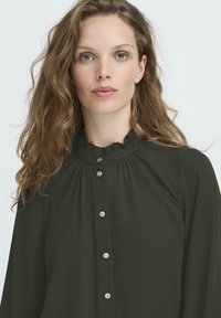 Jeune femme aux cheveux châtain clair ondulés, portant une blouse noire boutonnée avec un col à volants, faisant face à l'avant avec une expression neutre.