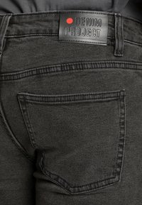 Pantalones vaqueros de mezclilla negra con una etiqueta de cuero con "DENIM PROJECT" grabada y un punto rojo, diseñados con un detalle en el bolsillo trasero.