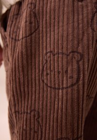 Tissu en velours côtelé brun présentant des nervures verticales et de subtils motifs de visage d'ours. La texture semble douce et veloutée.