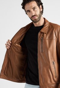 Man met een baard en krullend haar, gekleed in een zwart shirt en lichtblauwe spijkerbroek, die een bruine leren jas openhoudt om de binnenkant te tonen.