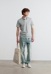 Jeune homme en polo gris et jean clair tenant un sac fourre-tout beige, debout sur un sol en béton contre un mur blanc uni.