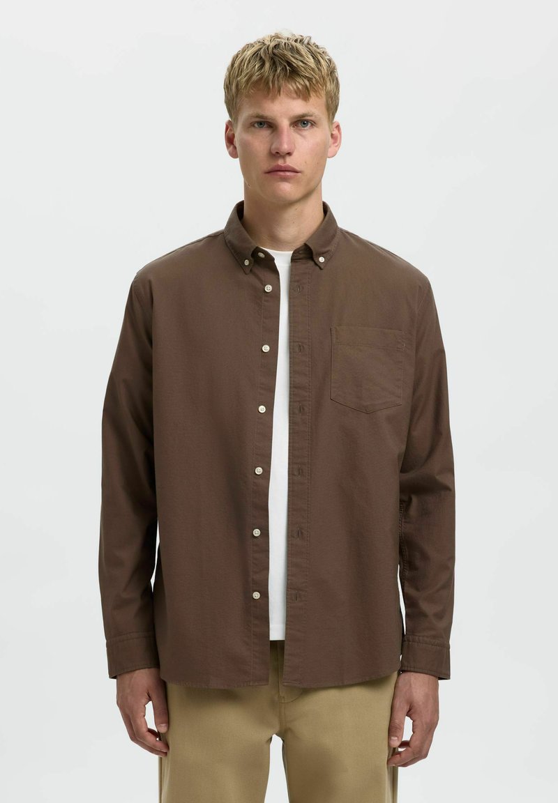 Camisa de botones marrón hecha de algodón, con un único bolsillo en el pecho, mangas largas y cuello. Lleva puesta una camiseta blanca debajo.