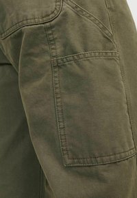 Gros plan d’un pantalon en tissu vert olive montrant une grande poche latérale rectangulaire avec des coutures visibles et une texture résistante.