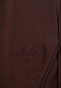 Dunkelbrauner Stoff mit eingesticktem Text "smilodox" in passendem Garn, zeigt feine Strickstruktur und Nahtdetail auf der rechten Seite.