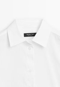 Chemise blanche habillée avec col pointu et deux boutons blancs visibles, montrant une étiquette noire à l'intérieur du col portant la mention Massimo Dutti, fabriquée au Maroc.