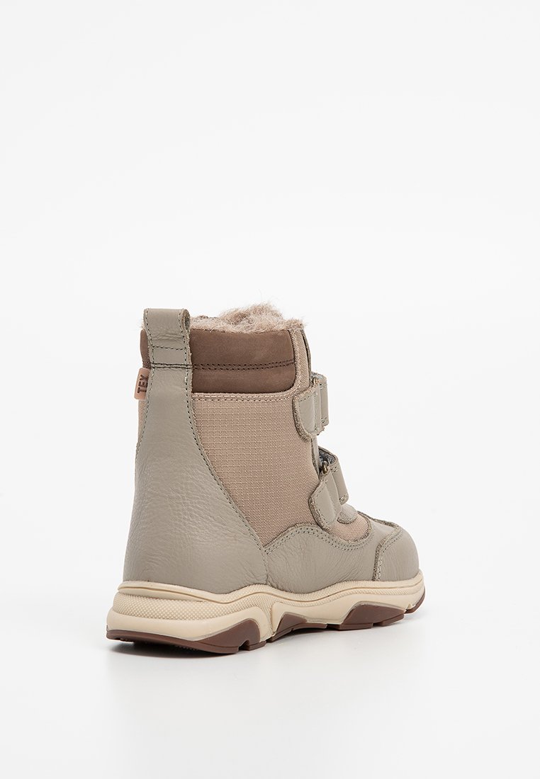 Beige syntetiskt boot med texturerade tygpaneler, två justerbara remmar och en mjuk imiterad pälsfoder vid kragen. Gummisulor ger bra grepp.