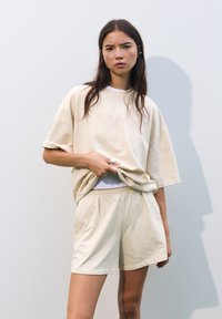 Beige oversize t-paita ja siihen sopivat shortsit pehmeästä kankaasta, rento istuvuus ja hienovarainen tekstuuri. Malli poseeraa kädet shortseissa.