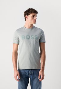 Camiseta de algodón verde claro con un gran logo "BOSS" central en el mismo color. Con mangas cortas y escote redondo. Llevada con jeans azules.