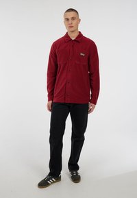 Jonge man die recht vooruit staat, gekleed in een rode fleece jack met knopen, zwarte broek en grijze sneakers met witte strepen.