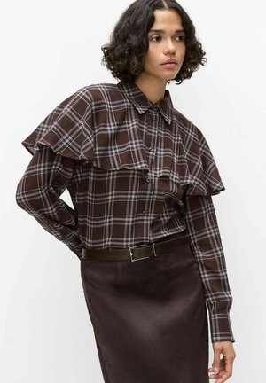 Femme aux cheveux courts et bouclés portant une chemise marron à carreaux boutonnée avec un rabat style cape et une jupe marron foncé avec ceinture.