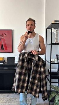 Man in white tank top holding plaid jacket on hanger in one hand, applying makeup brush to face indoors with art and shelves in background.