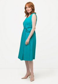 Vestito smanicato color turchese con vita annodata, lunghezza al ginocchio, realizzato in tessuto morbido. Indossato con scarpe con tacco beige, caratterizzate da un motivo testurizzato.
