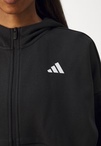 Čierna mikina na zips vyrobená z jemného materiálu, s výrazným bielym logom Adidas na hrudi a klasickým strihom.
