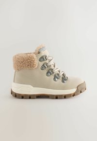 Beige vinter vandrestøvle med fleecekrave, metaløjer, hvide snørebånd og tyk, grov brun sål, set fra siden mod en lys baggrund.