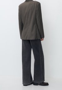 Harmaa houndstooth-bleiseri, jossa on muotoiltu istuvuus, yhdistettynä leveälahkeisiin harmaisiin denim-housuihin. Bleiserissä on viistetty kaulus ja sileä pinta.