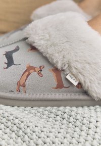Grauer Hausschuh mit einem flauschigen, hellgrauen Futter und einem Dackelmuster. Etikett mit der Aufschrift "NEXT". Verfügt über eine strukturierte, weiche Stoffoberfläche.