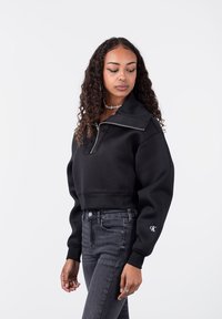 Sudadera negra corta con cuello alto y cremallera, con puños acanalados. Combinada con jeans ajustados de color gris oscuro. Detalle de logo minimalista.