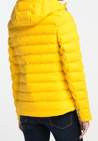 Gul puffjacka med kviltat mönster, huva och elastiska ärmmuddar. Slät, glansig textur och synliga sömnadsdetaljer.