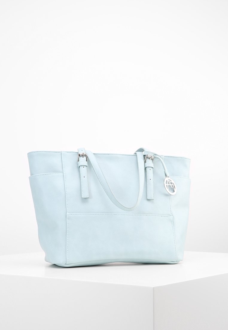 Sac tote en cuir bleu clair avec des bretelles à double boucle et un charm logo rond argenté, présenté sur un piédestal blanc sur fond uni.