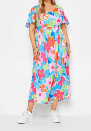 Femme portant une robe maxi fleurie colorée avec des manches volantées et des baskets blanches, debout devant un fond uni.