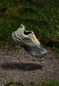 Lightweight gray and green sneaker with yellow laces, mesh upper, textured accents, and rubber sole, floating above a gravel surface.