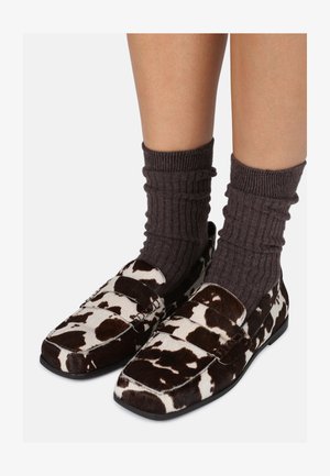 Brūnās un baltās govs druku loafers, kas izgatavoti no teksturētām materiāliem, ar plakanu zoli un strukturētu dizainu, kombinēti ar tumšiem ribotajiem zeķēm.