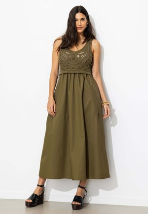 Femme portant une robe sans manches vert olive avec un haut en crochet, des sandales à plateforme noires et des bracelets dorés, debout contre un mur blanc.