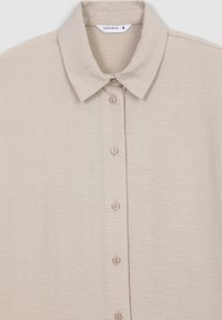 Camicia beige con bottoni, realizzata in un tessuto morbido e testurizzato. Presenta un colletto e cinque bottoni coordinati lungo la parte anteriore. L'etichetta indica la taglia small.