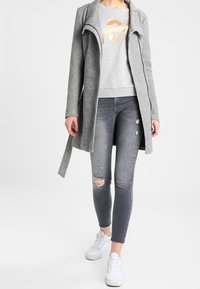 Abrigo largo gris claro con cuello de chal, usado sobre una sudadera gris con un gráfico dorado, combinado con jeans oscuros desgastados y zapatillas blancas.